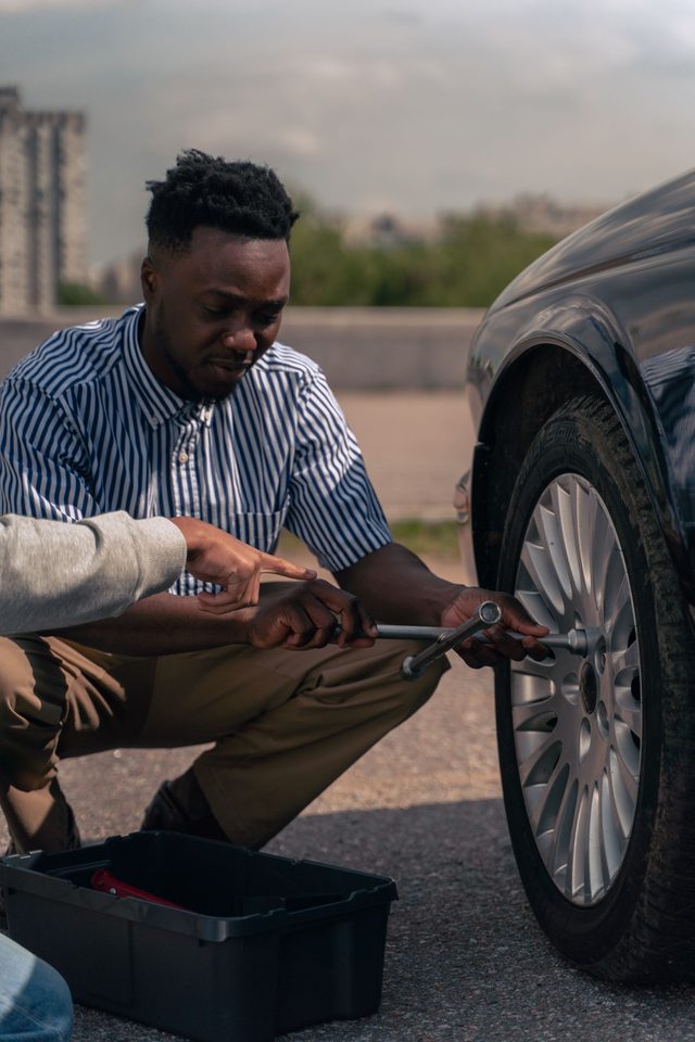 Two people changing a wheel at the roadside with a breaker bar and a tool tray